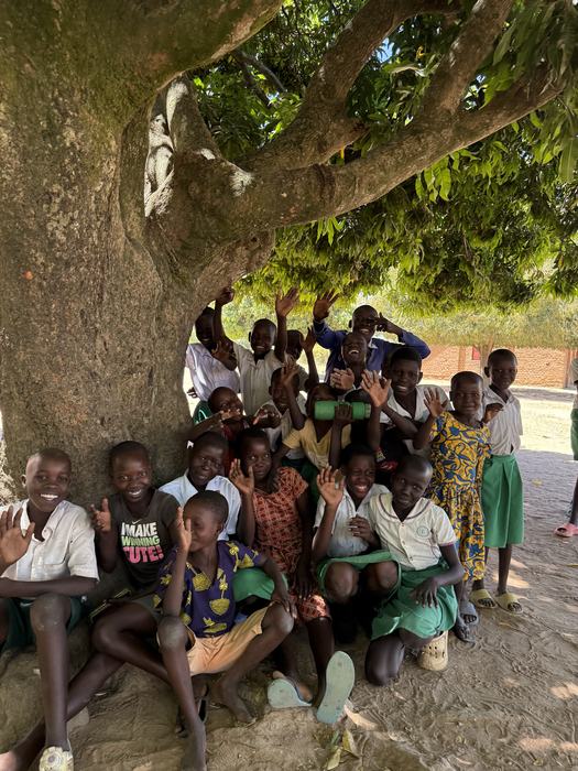Children waving and smiling under a mango tree